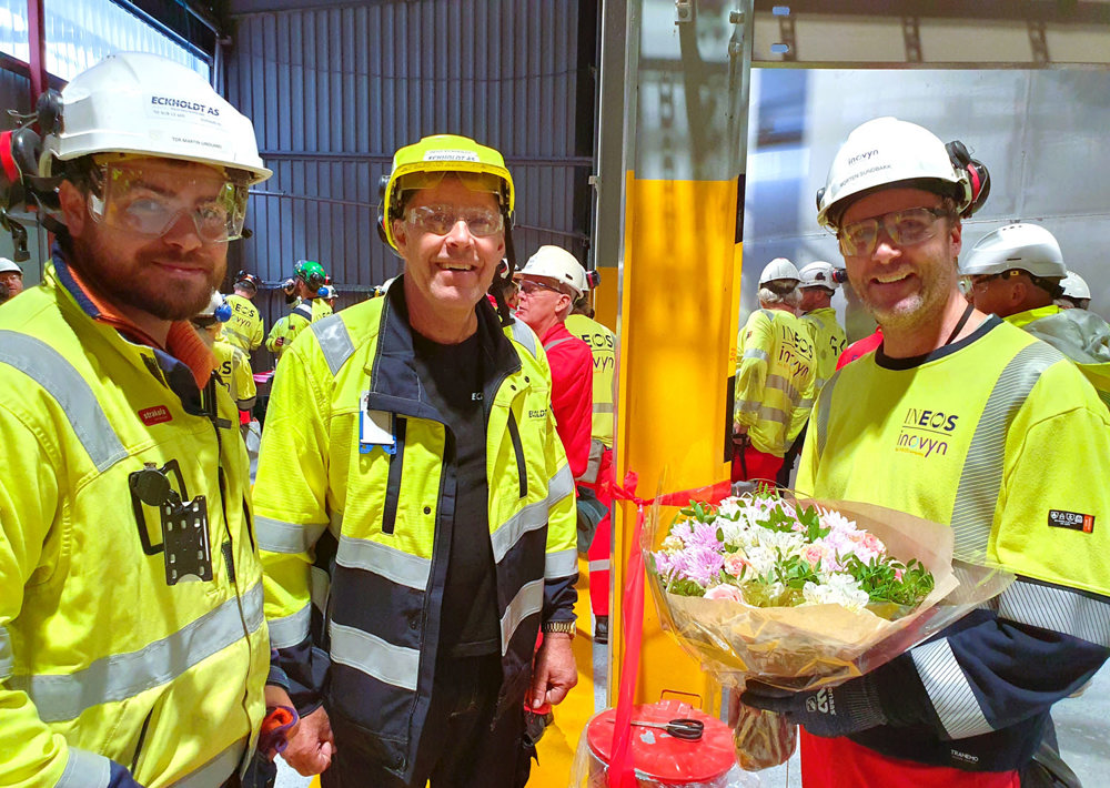 three men standing, posing, one man holding flowers, production hall
