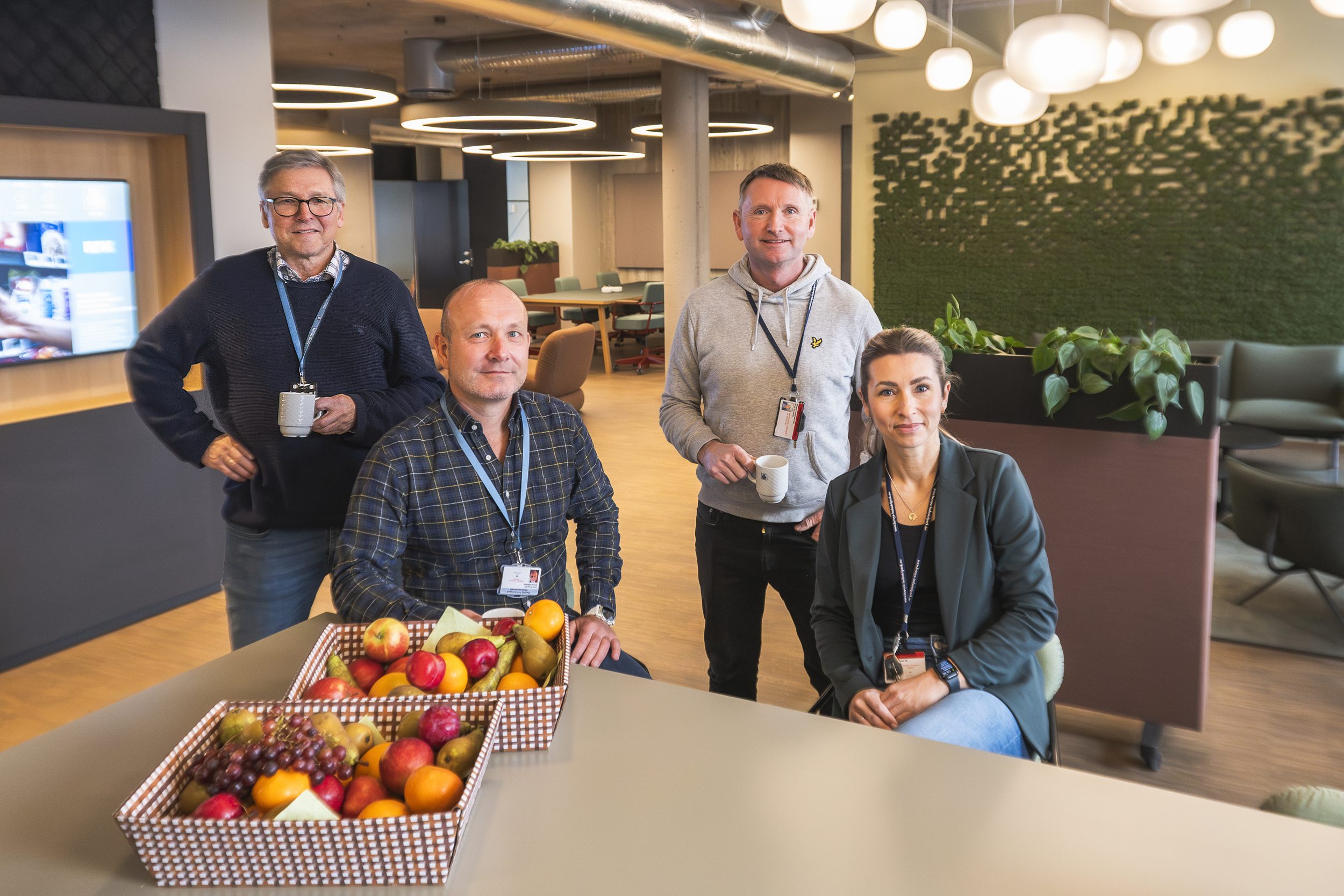 office environment, four persons, posing, standing and sitting by a high table. Fruit on the table, green moss on the wall behind. Furniture in background.