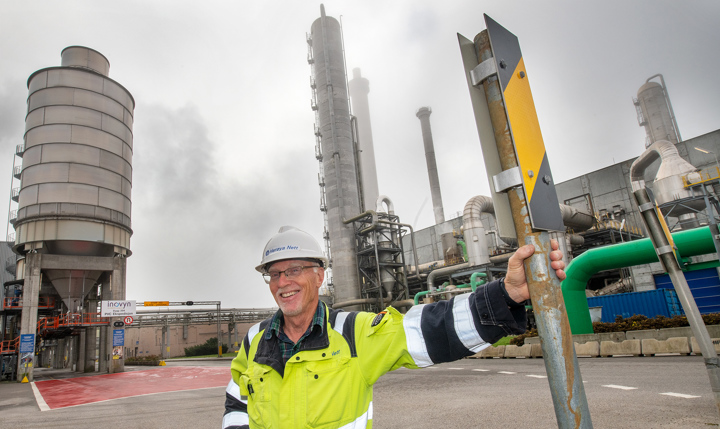 mann poserer, gul jakke, hvit hjelm, strak venstre hånd ut og holder i et skilt, industrianlegg i bakgrunnen