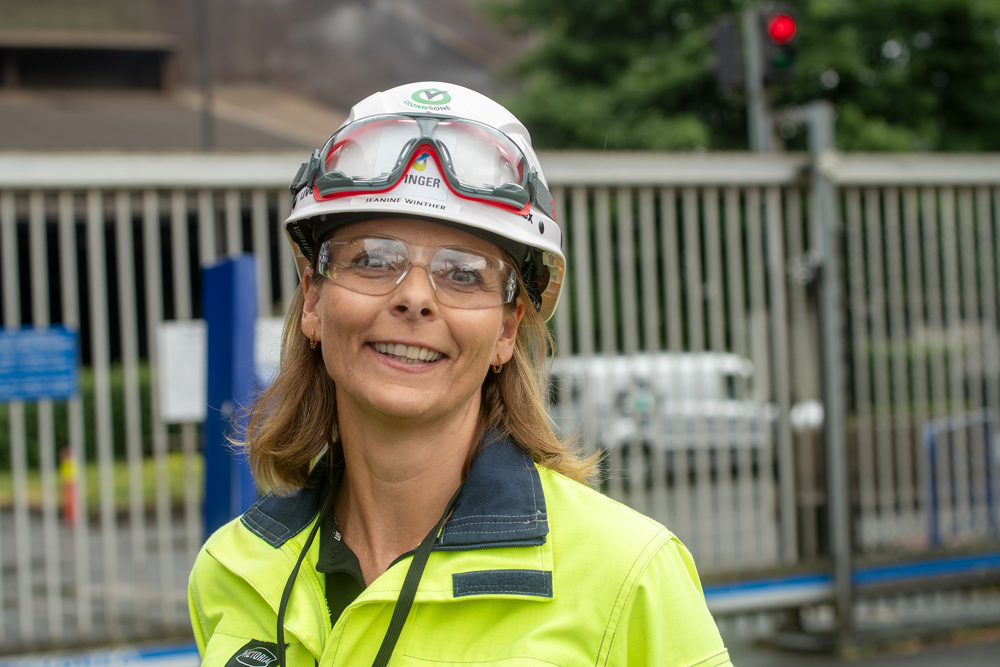 portrait of woman, goggles, white helmet, yellow and blue jacket, standing outside industrial fencing.