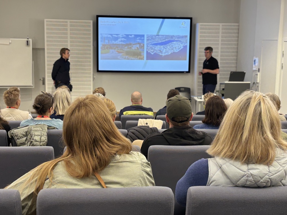two persons in front presenting for audience sitting in chairs in an auditorium.