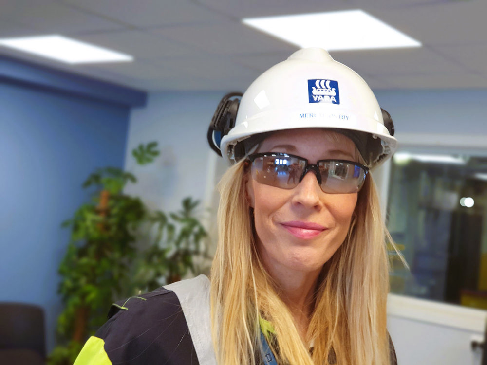 portrait of young woman with long blond hair, wearing white helmet and dark glasses, standing in a meeting room.