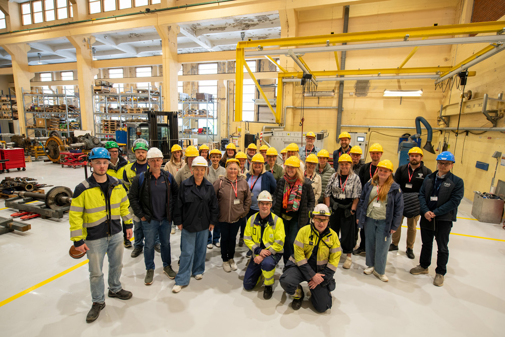 large group of people standing in a work shop with yellow helmets, two people sitting down in front row.