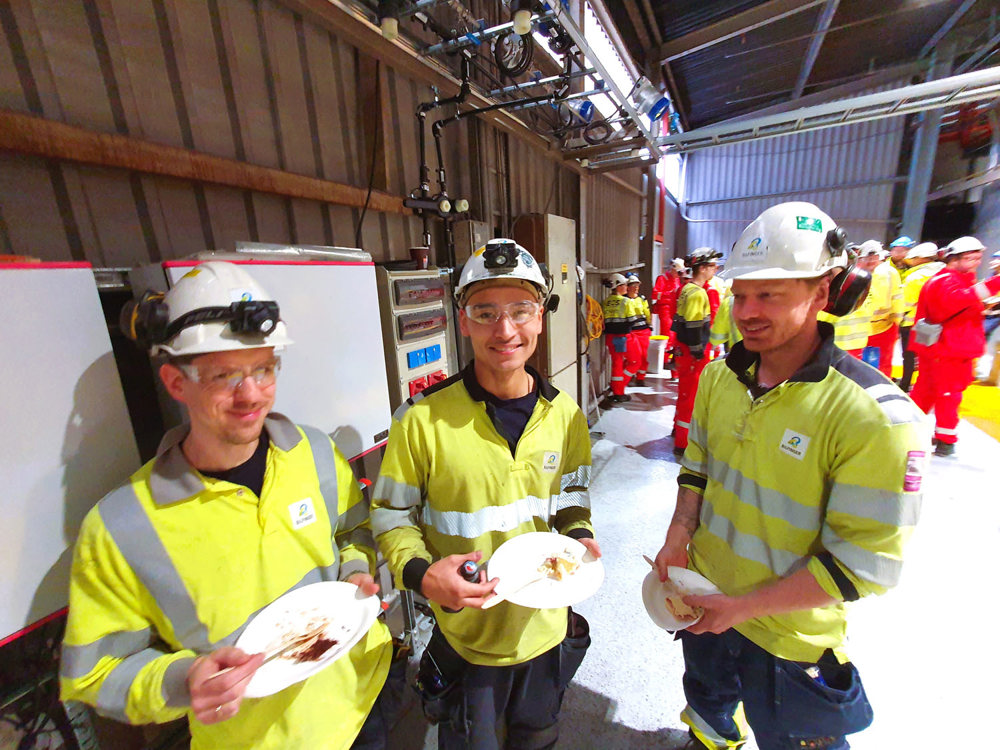three young men, posing, standing eating cake, wearing yellow jumpers and blue trousers and white helmets, production hall