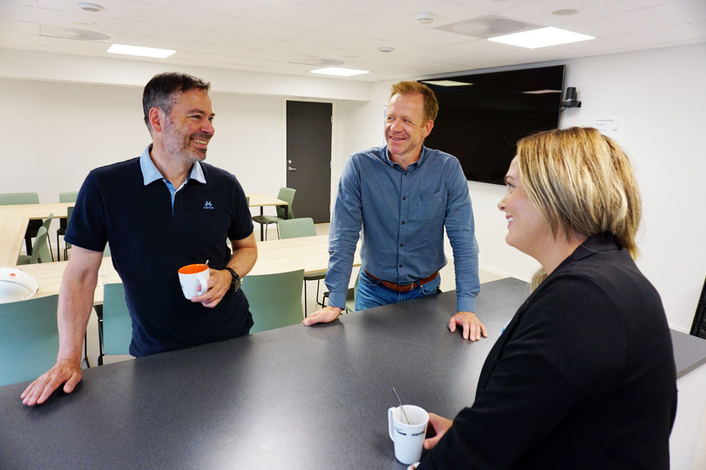 three people standing at a high table in a canteen, green chairs black table top