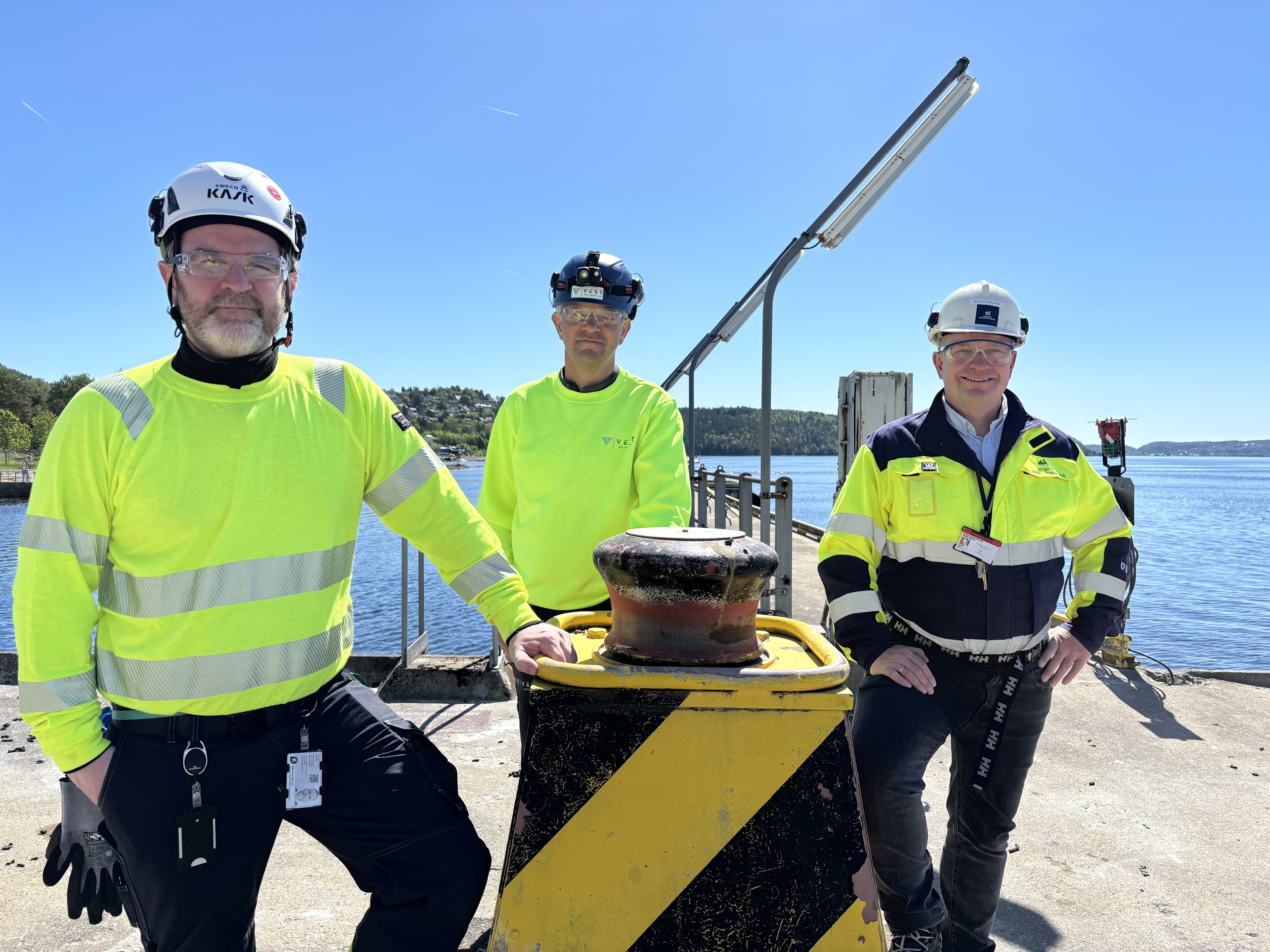 tre menn i verneutstyr, står rundt en pullert på kai, poserer