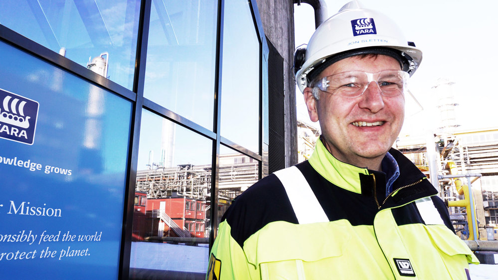 portrait of man, smiling, posing, white helmet, yellow jacket, logo on glass wall behind him