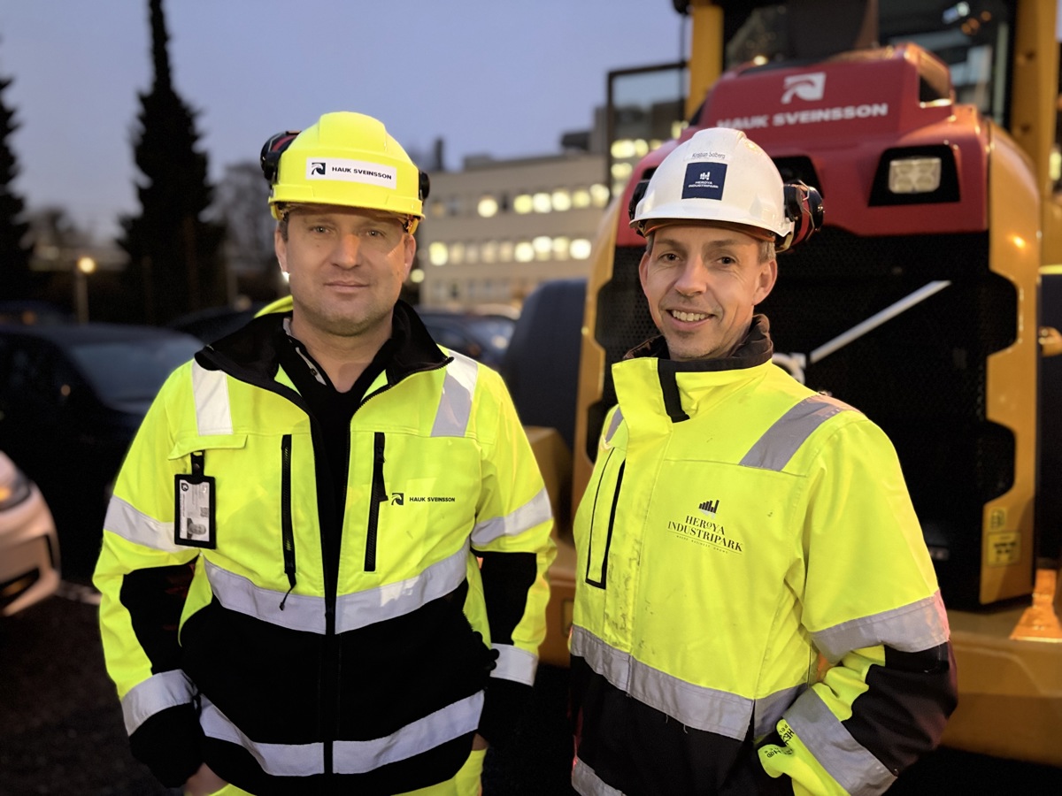 to menn i hjelm og gul jakke, poserer, hjullaster i bakgrunnen