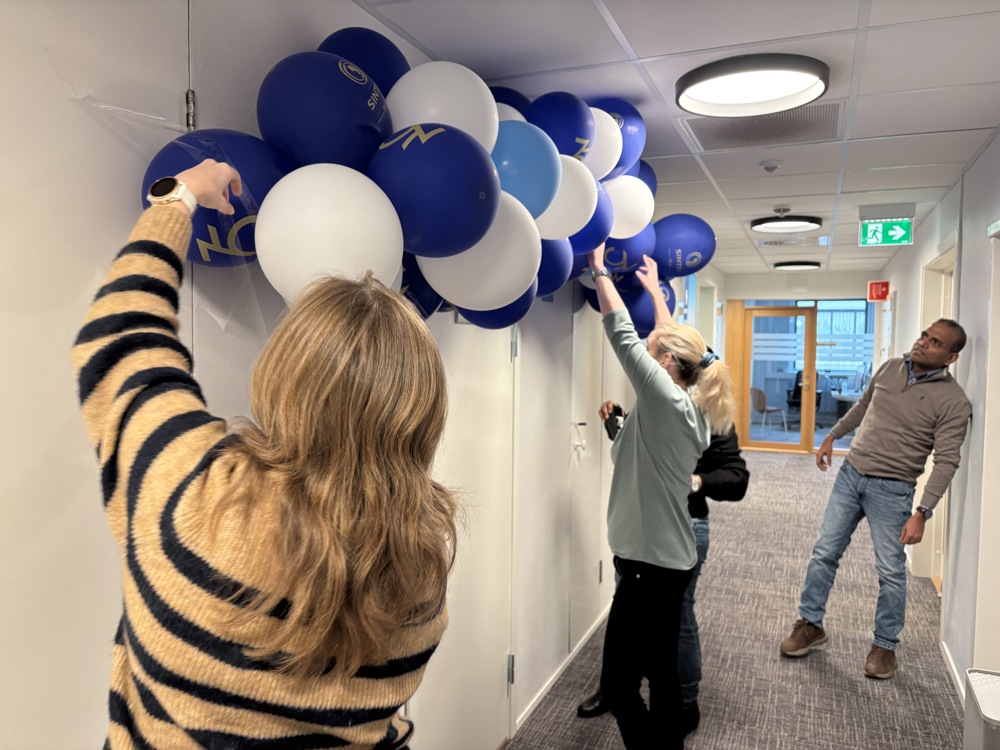 tre personer henger opp rad med ballonger oppunder taket i kontorlokalet