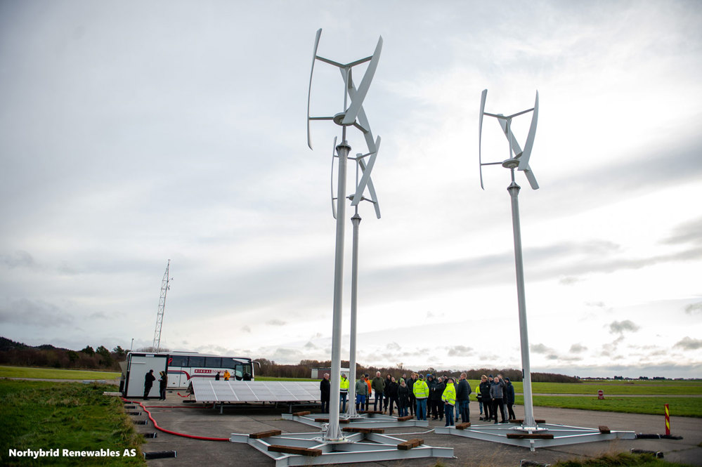 three wind mills, vertical blades, solar panels, people gathering on the site.