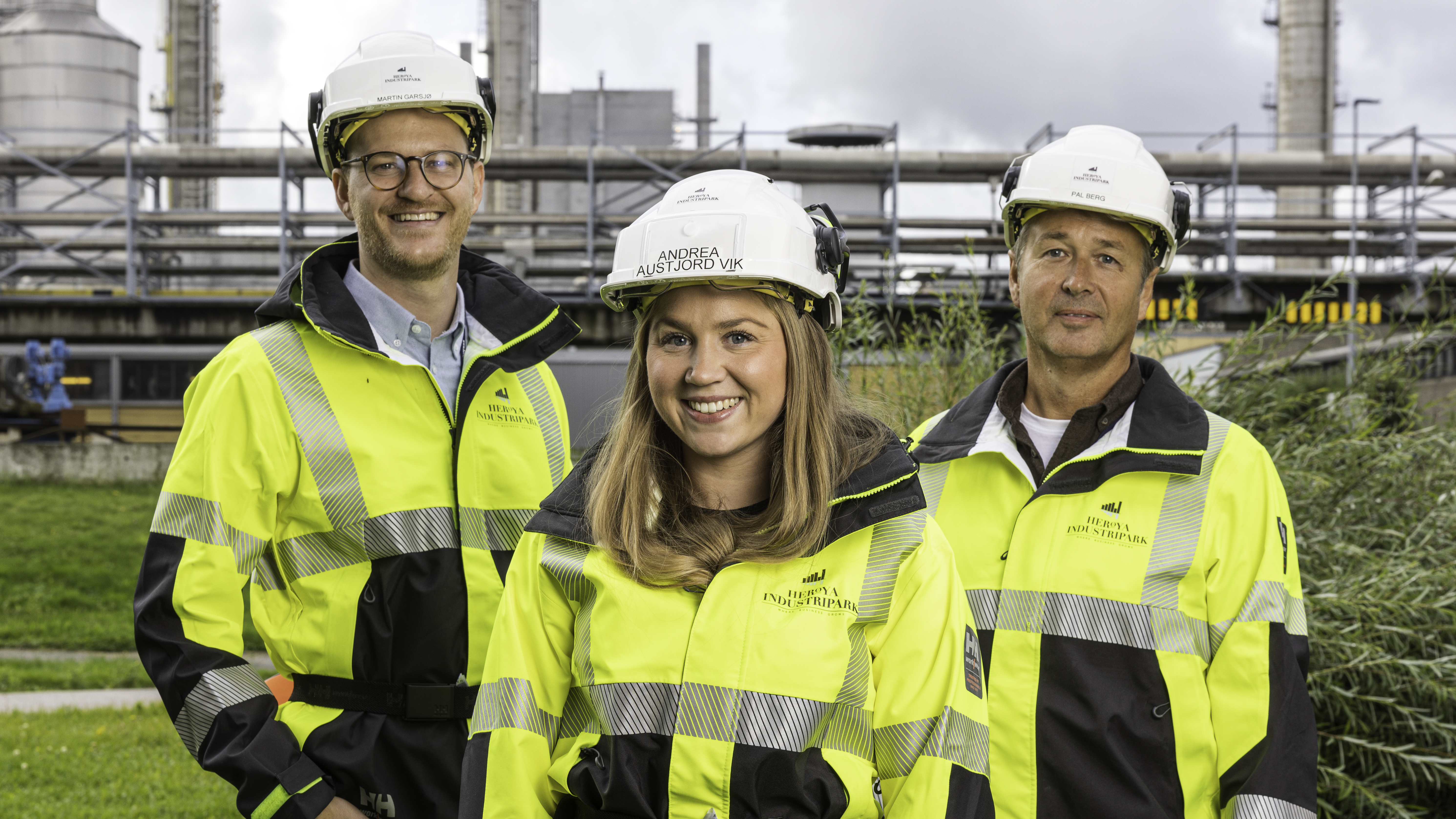 tre personer, poserer, verneutstyr, gule jakker, hvite hjelmer, ute i industripark, bakgrunn prosessindustri, grøntanlegg.