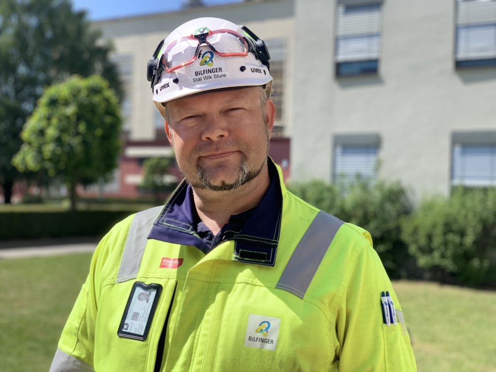 man posing, wearing helmet and yellow working clothes, portrait, trees and lawn in background and building with windows.