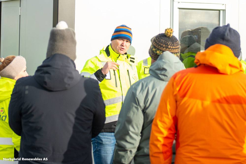 Man stands close to building and talks to people in front of him, all wearing thick winter coats and hoods.