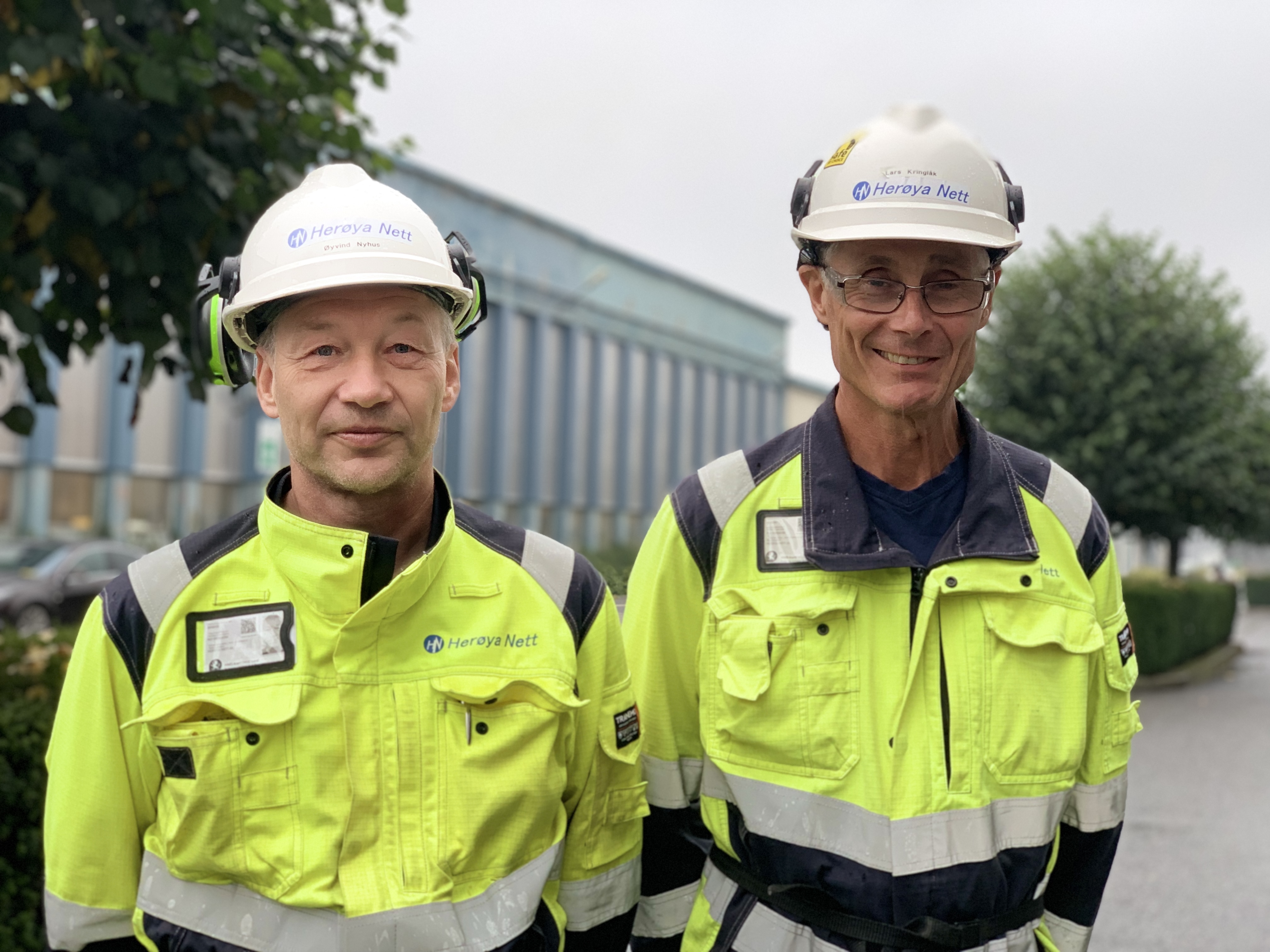 two men next to eachother, posing, yellow jackets, white helmets, in a street in an industry area, green trees and an indstrial building in background