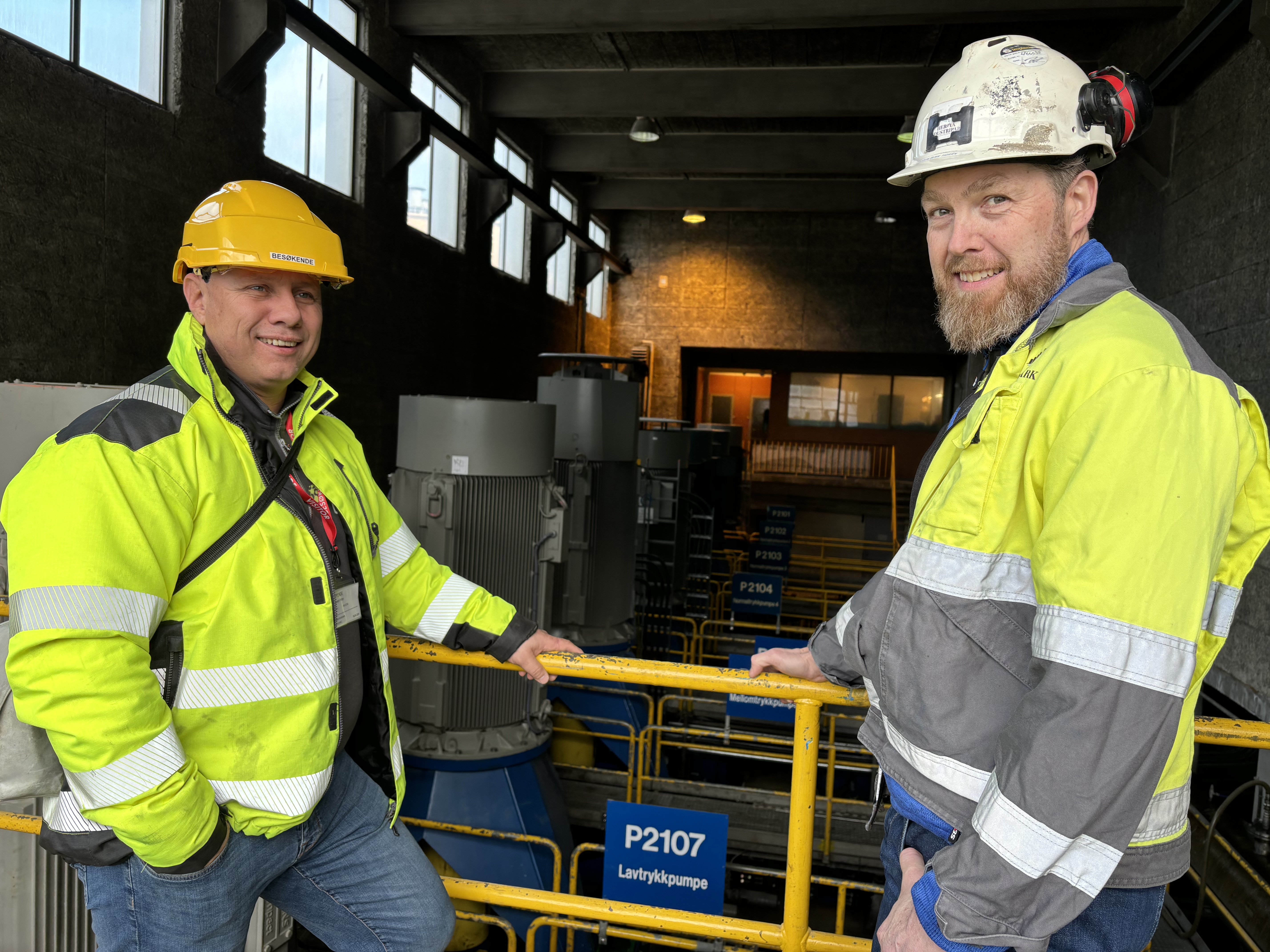two men pose and stand by a yellow railing, protective equipment, below is a pumping station, large raw water pumps, blue signs on each pump.