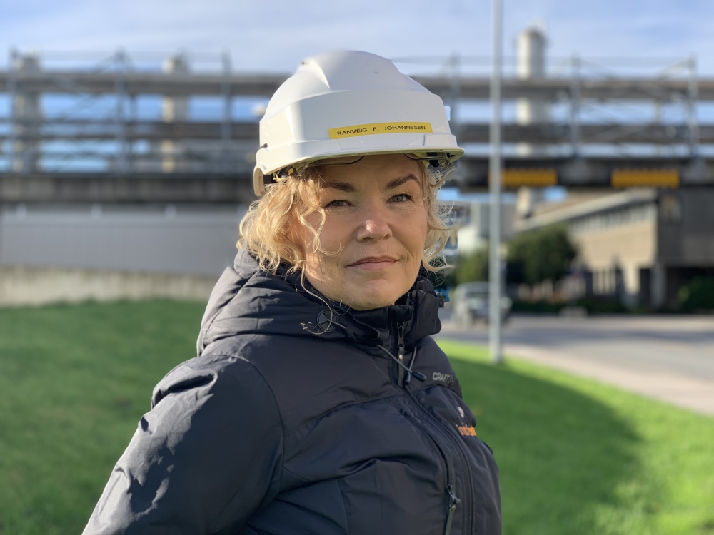 portrett av kvinne, blå jakke, hvit hjelm, poserer, industripark i bakgrunnen