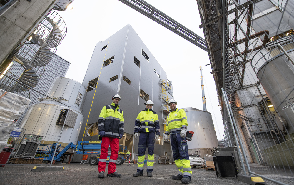 Tre personer står på bakken foran bygg og stor kran.