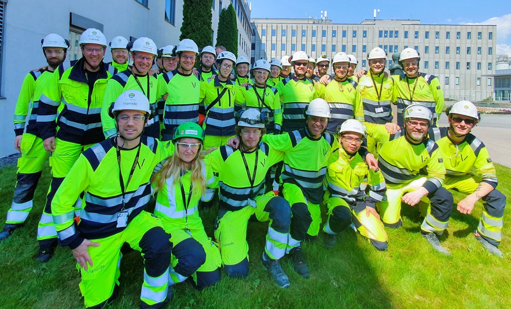 Team photo, one line standing, one line kneeling, all wearing yellow and blue working clothes, white helmets, background green area, outside office building.